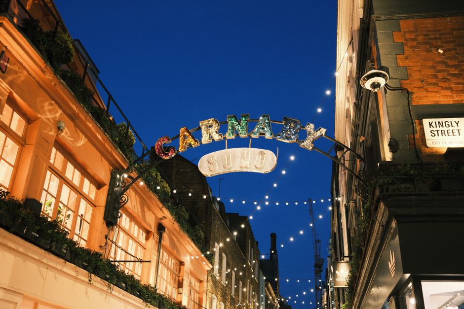 Nighttime photograph of Carnaby Street in Soho, London, with a prominent illuminated archway featuring the colorful “CARNABY” sign and a smaller, white “SU14” sign hanging below. The arch span