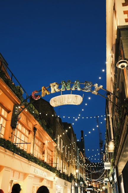 Nighttime photograph of Carnaby Street in Soho, London, with a prominent illuminated archway featuring the colorful “CARNABY” sign and a smaller, white “SU14” sign hanging below. The arch span