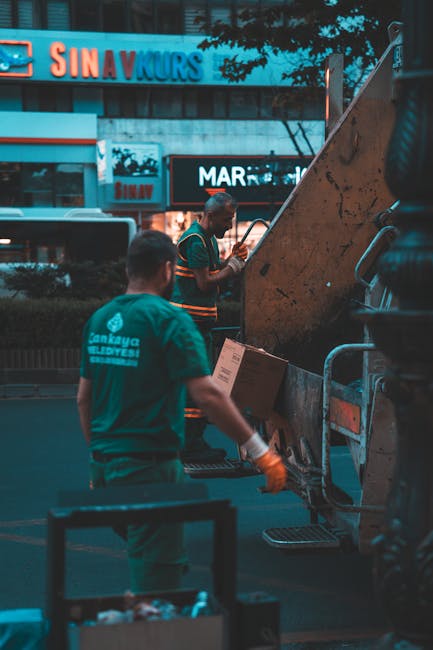 A narrow urban alleyway cluttered with discarded rubbish and waste materials, featuring a large, wheeled trash bag made of coarse, woven fabric in the foreground, positioned centrally. Behind it, ther