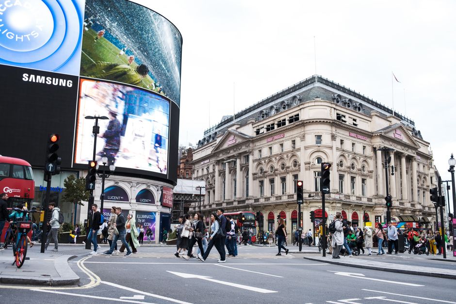 A nighttime scene of Piccadilly Circus in London showing illuminated digital billboards and advertisements on large electronic screens mounted on the building facades. In the foreground, a black metal